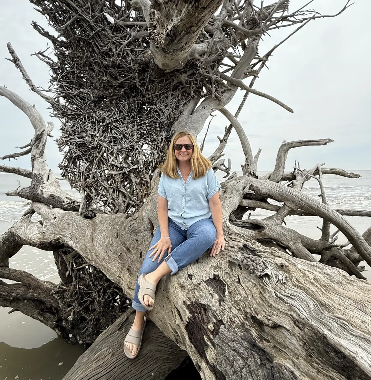 Andrea sitting on a large piece of driftwood on a beach, smiling, with the sea behind her.